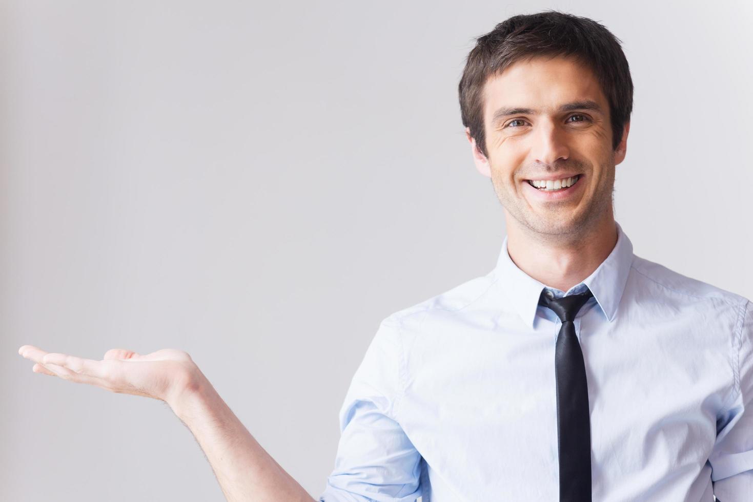 zakenman Holding kopiëren ruimte. gelukkig jong Mens in overhemd en stropdas Holding kopiëren ruimte Aan zijn hand- en glimlachen terwijl staand tegen grijs achtergrond foto