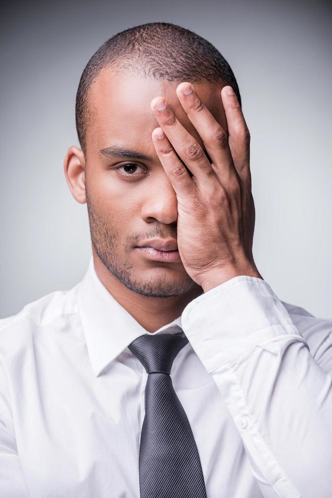 gevoel moe en depressief. depressief jong Mens in overhemd en stropdas aan het bedekken een deel van zijn gezicht met hand- en op zoek Bij camera terwijl staand tegen grijs achtergrond foto