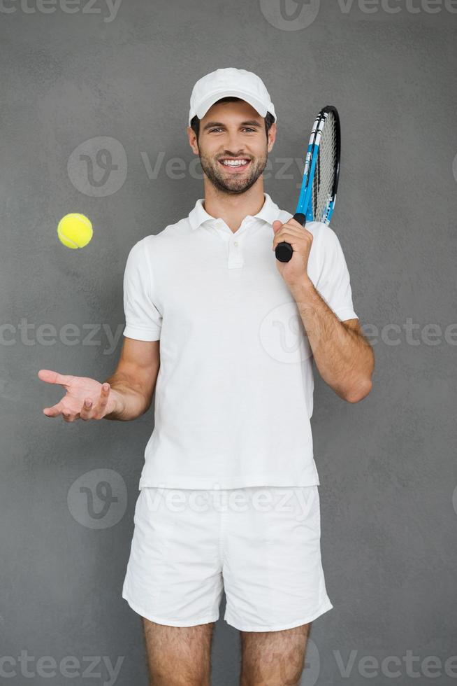 klaar naar Speel. vrolijk jong Mens in sport- kleren draag- tennis racket Aan zijn schouder en het werpen tennis bal terwijl staand tegen grijs achtergrond foto