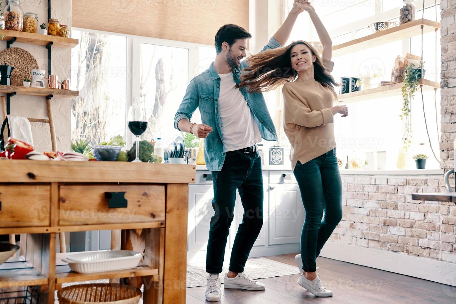 twee harten gevulde met liefde. vol lengte van mooi jong paar in gewoontjes kleding dansen en glimlachen terwijl staand in de keuken Bij huis foto