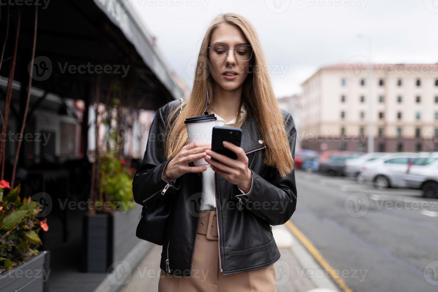 jong vrouw typen een bericht Aan een mobiel telefoon met een kop van koffie in haar handen tegen de backdrop van de stedelijk landschap De volgende naar een cafe foto