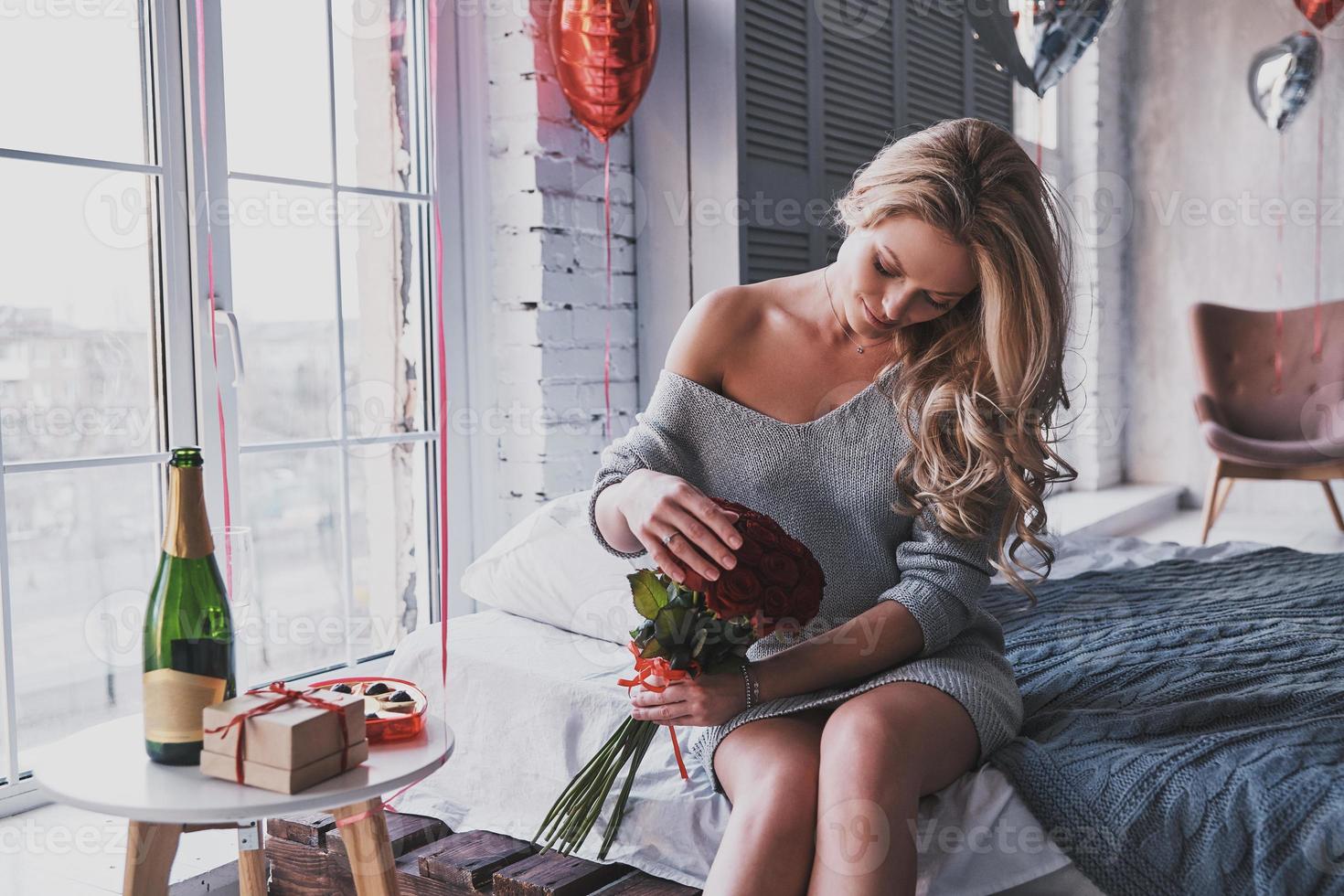 haar favoriete bloemen. aantrekkelijk jong vrouw Holding een boeket van rood rozen en glimlachen terwijl zittend Aan de bed Bij huis foto