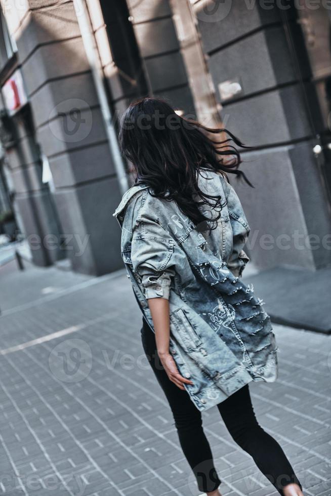 in een haast je. achterzijde visie van jong vrouw in denim jasje wandelen naar beneden de straat foto
