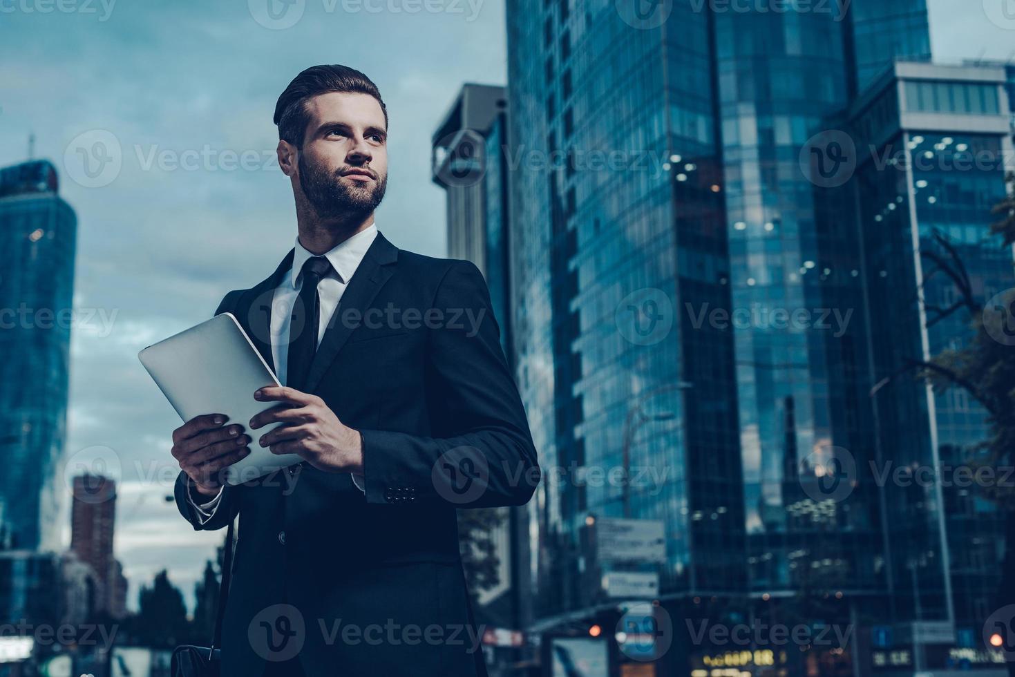 deze is mijn stad. nacht tijd beeld van zelfverzekerd jong Mens in vol pak Holding digitaal tablet en op zoek weg terwijl staand buitenshuis met stadsgezicht in de achtergrond foto