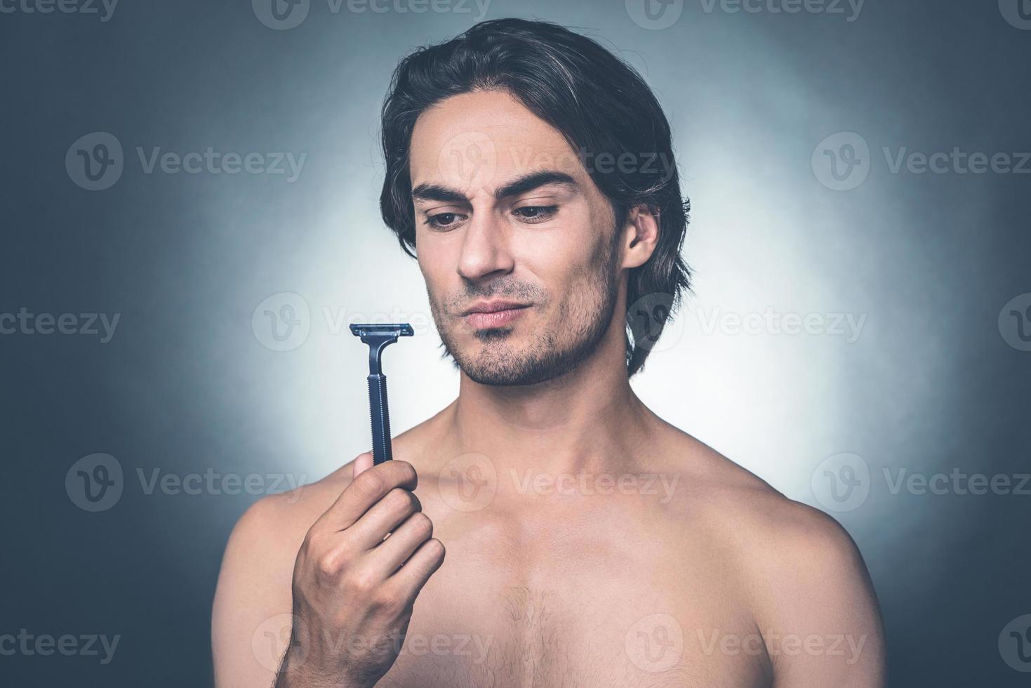 werkelijk slecht scheermes. portret van attent jong zonder shirt Mens op zoek Bij scheermes terwijl staand tegen grijs achtergrond foto