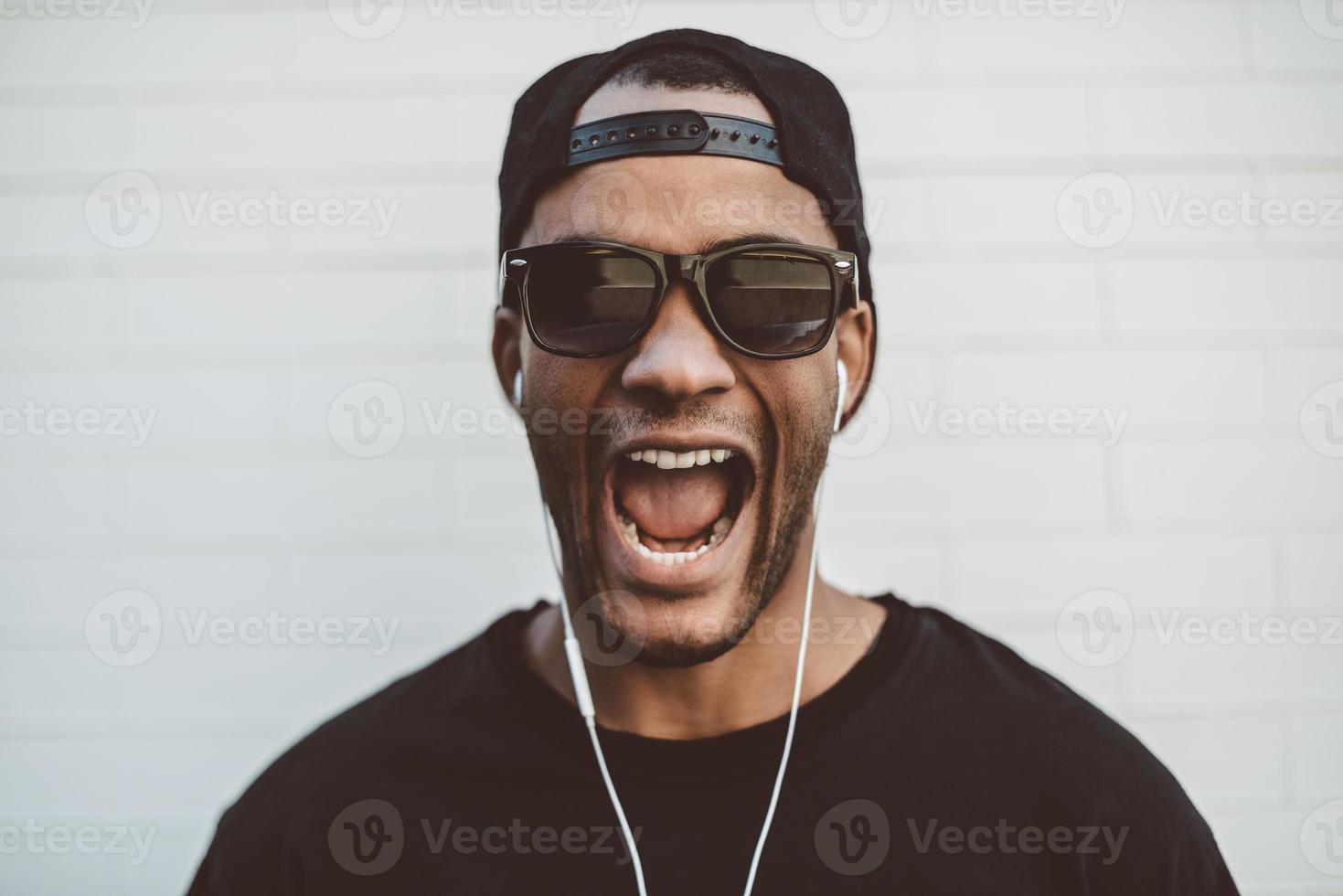 genieten van zijn favoriete muziek. knap jong Afrikaanse Mens in hoofdtelefoons op zoek Bij camera en houden mond Open terwijl staand in voorkant van de stoned muur buitenshuis foto