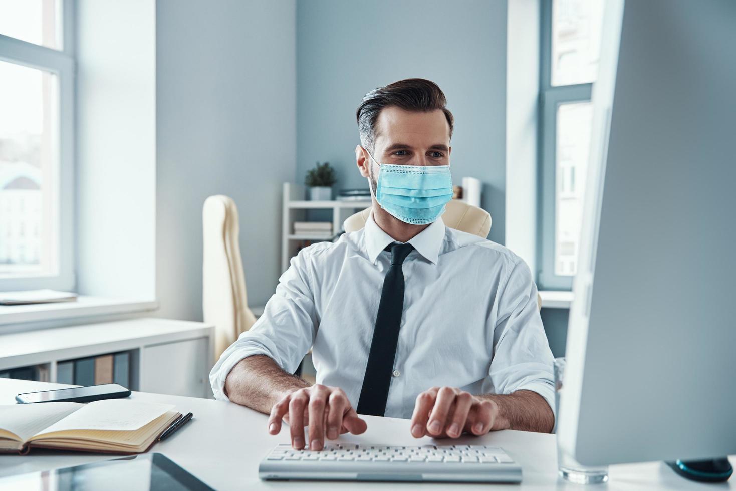 zelfverzekerd jong zakenman in beschermend gezicht masker werken Aan computer terwijl zittend in de kantoor foto