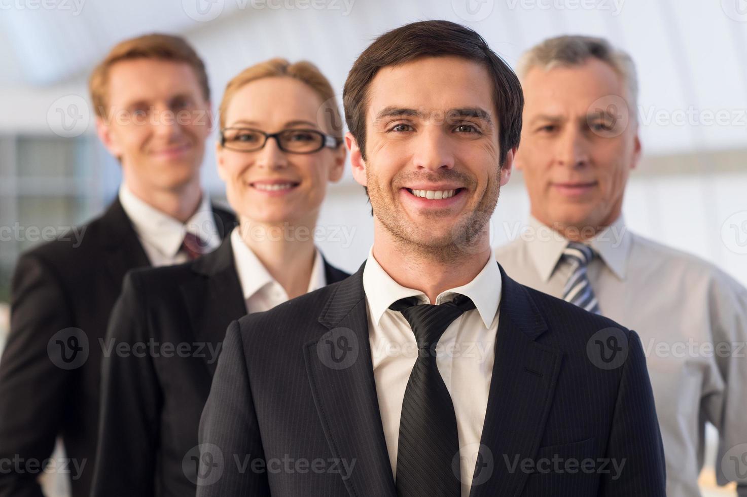 elke dag leider. zelfverzekerd jong Mens in formele kleding op zoek Bij camera en glimlachen terwijl zijn collega's staand achter hem foto