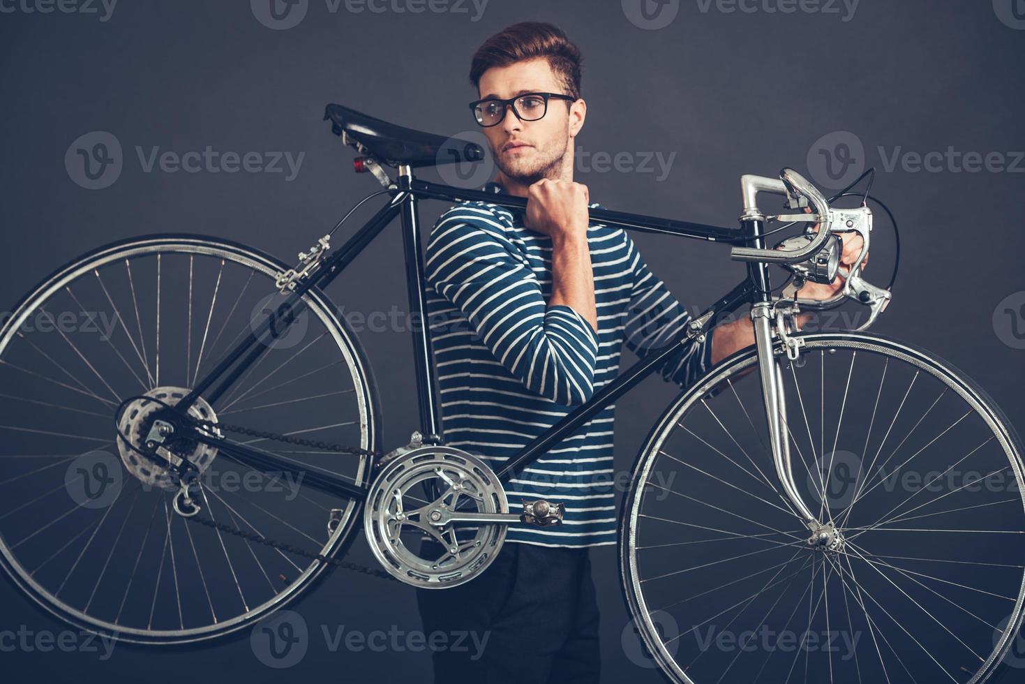 in zijn eigen stijl. zelfverzekerd jong knap Mens in bril draag- zijn retro gestileerd fiets Aan schouder en op zoek weg terwijl staand tegen grijs achtergrond foto