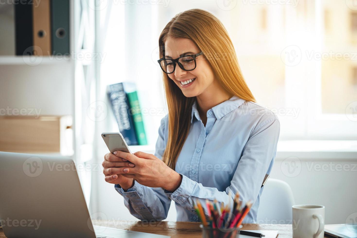 Super goed bericht vrolijk jong mooi vrouw in bril gebruik makend van haar smartphone met glimlach terwijl zittend Bij haar werken plaats foto