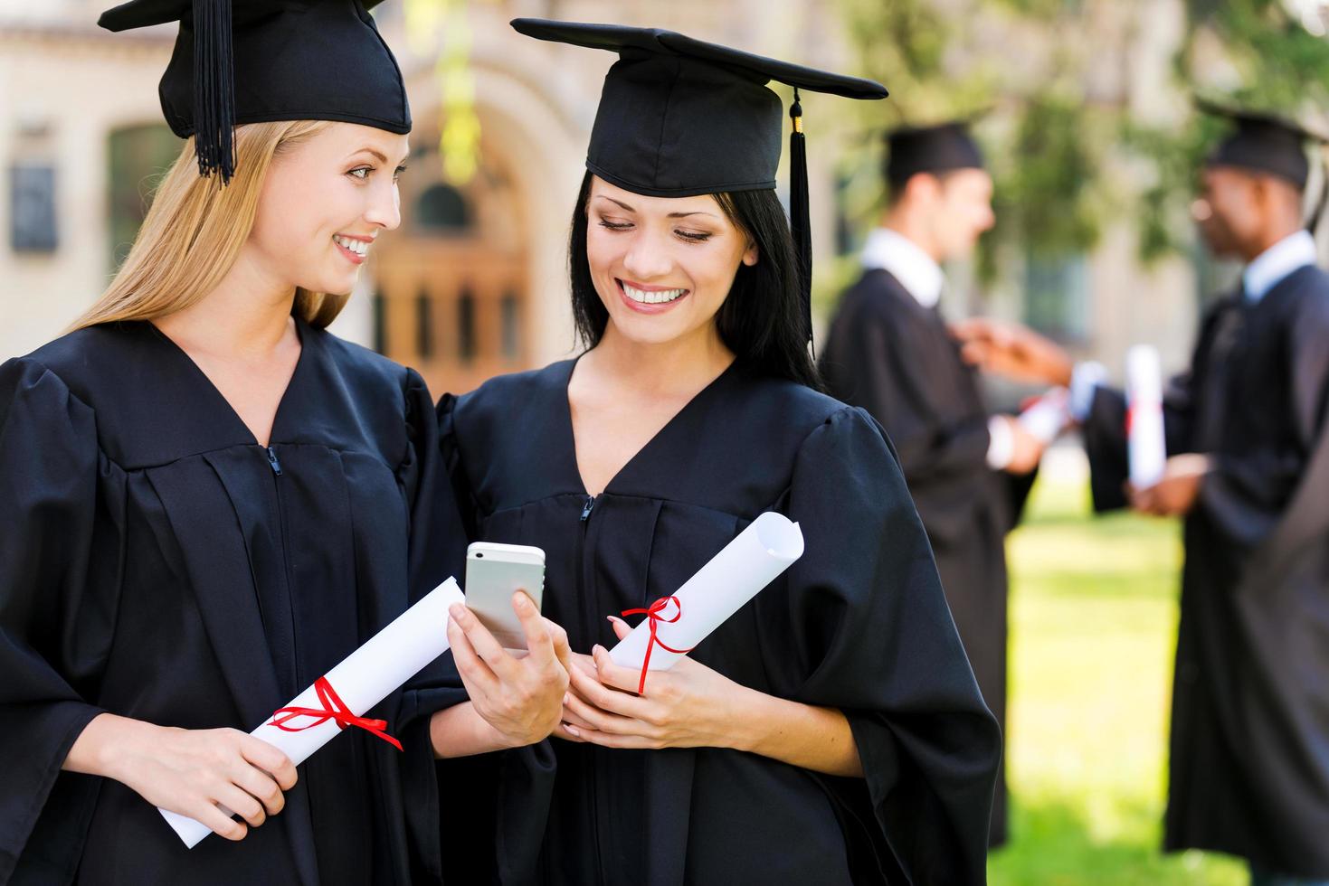 gelukkig afgestudeerden. twee gelukkig Dames in diploma uitreiking jurken op zoek Bij de mobiel telefoon samen en glimlachen terwijl twee mannen staand in de achtergrond foto