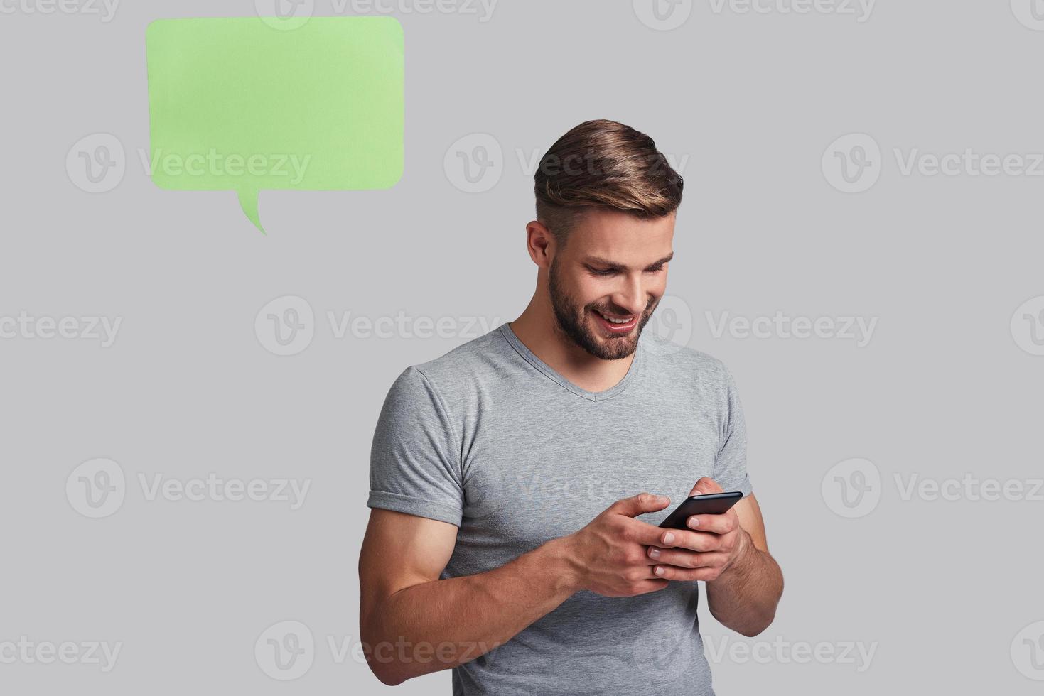 snel bericht naar vriend. knap jong glimlachen Mens Holding slim telefoon en op zoek Bij het terwijl staand tegen grijs achtergrond foto