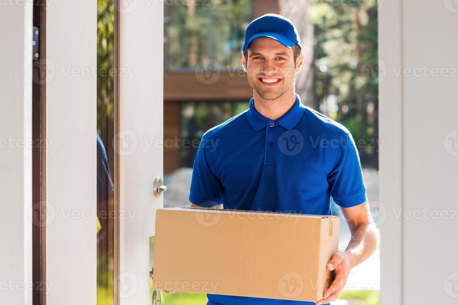 levering met glimlach. vrolijk jong koerier Holding een karton doos terwijl staand Bij de Ingang van appartement foto