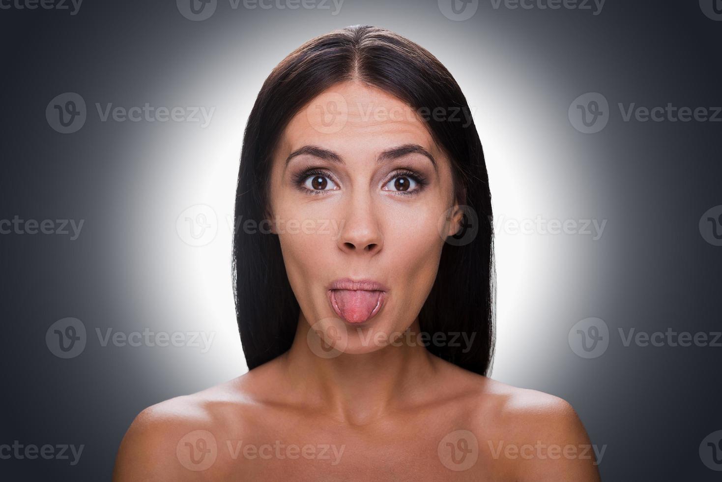 grappig grijns. portret van mooi jong zonder shirt vrouw op zoek Bij camera en tonen haar tong terwijl staand tegen grijs achtergrond foto