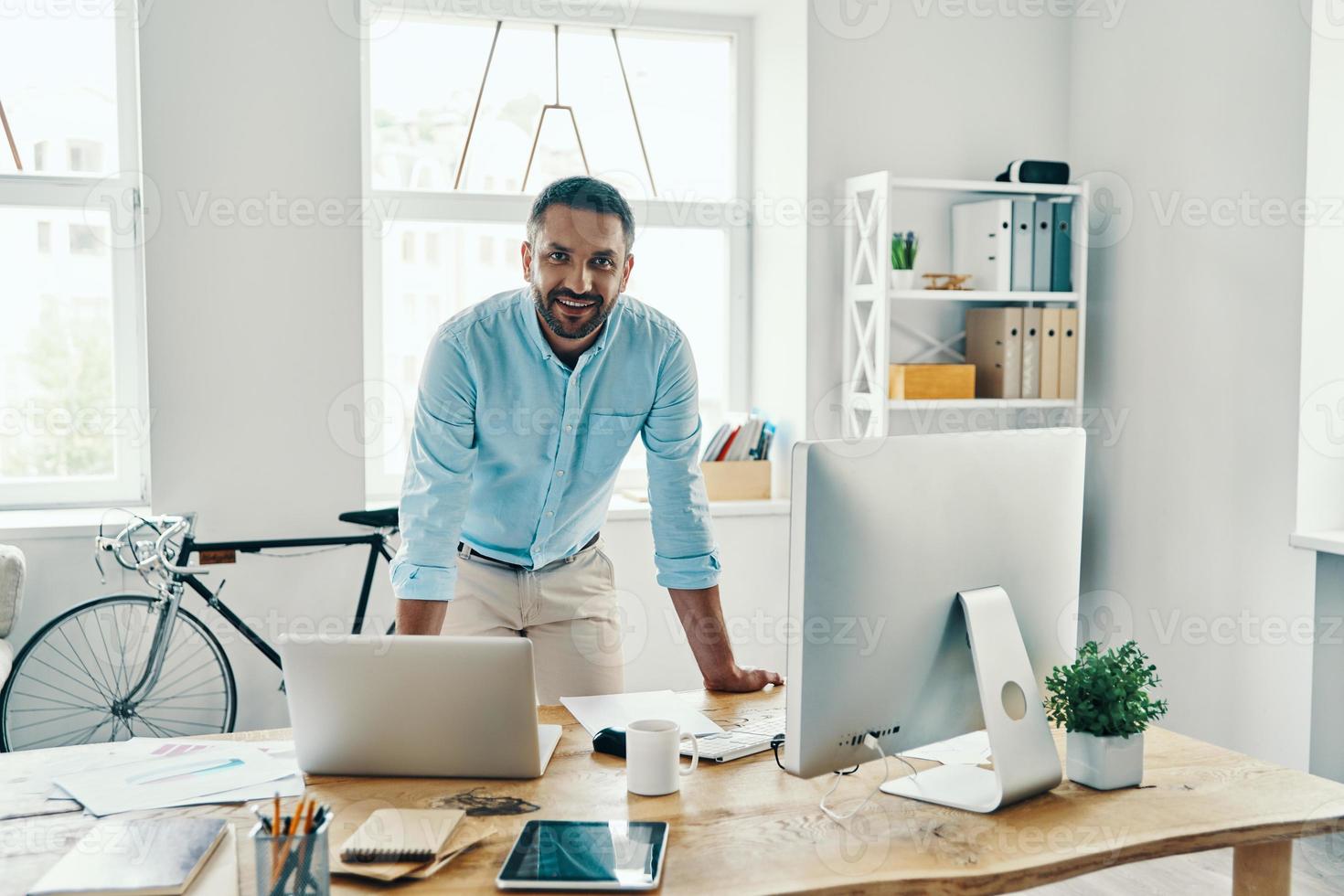 knap midden- leeftijd Mens in slim gewoontjes slijtage op zoek Bij camera en glimlachen terwijl staand in de kantoor foto