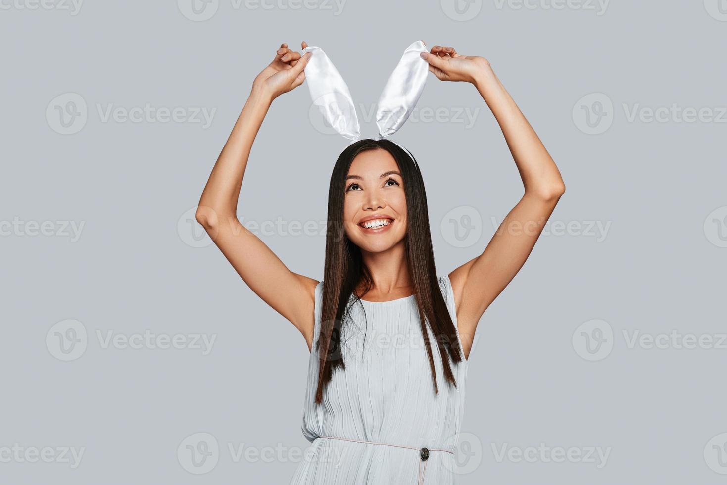 aan het doen wat dan ook ze wil. mooi jong Aziatisch vrouw aanraken haar konijn oren en glimlachen terwijl staand tegen grijs achtergrond foto