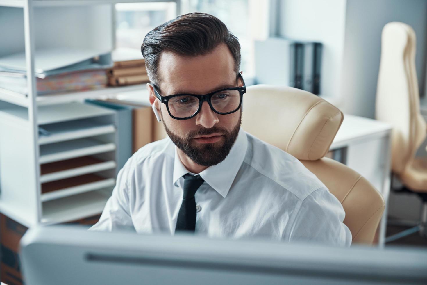 geconcentreerd jong Mens in overhemd en stropdas werken Aan computer terwijl zittend in de kantoor foto