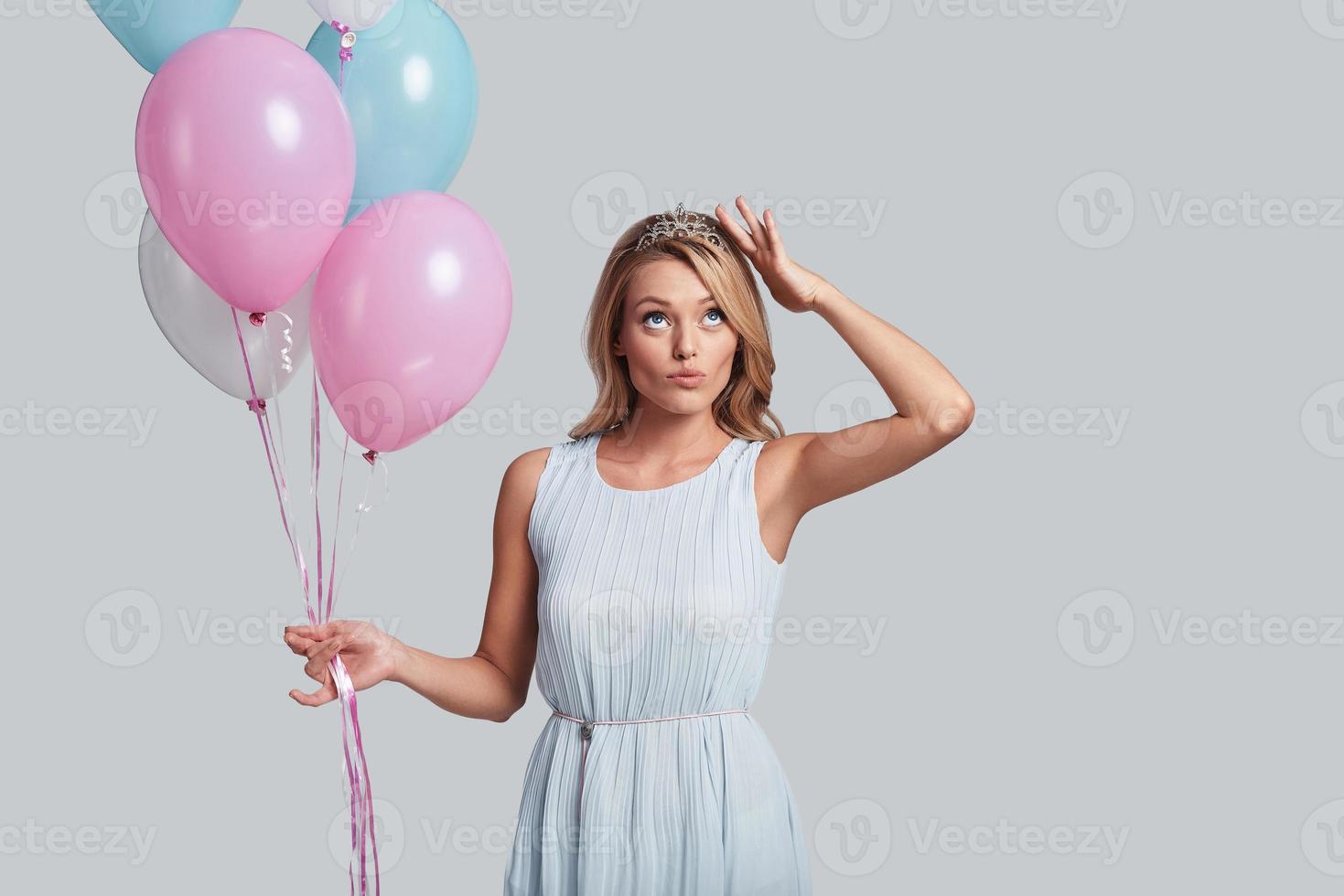 controle de kroon. aantrekkelijk jong vrouw Holding ballonnen en aanraken haar hoofd met hand- terwijl staand tegen grijs achtergrond foto