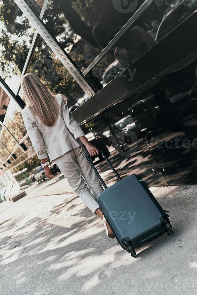 zelfverzekerd zakenvrouw. vol lengte achterzijde visie van jong vrouw in pak trekken bagage terwijl wandelen buitenshuis foto