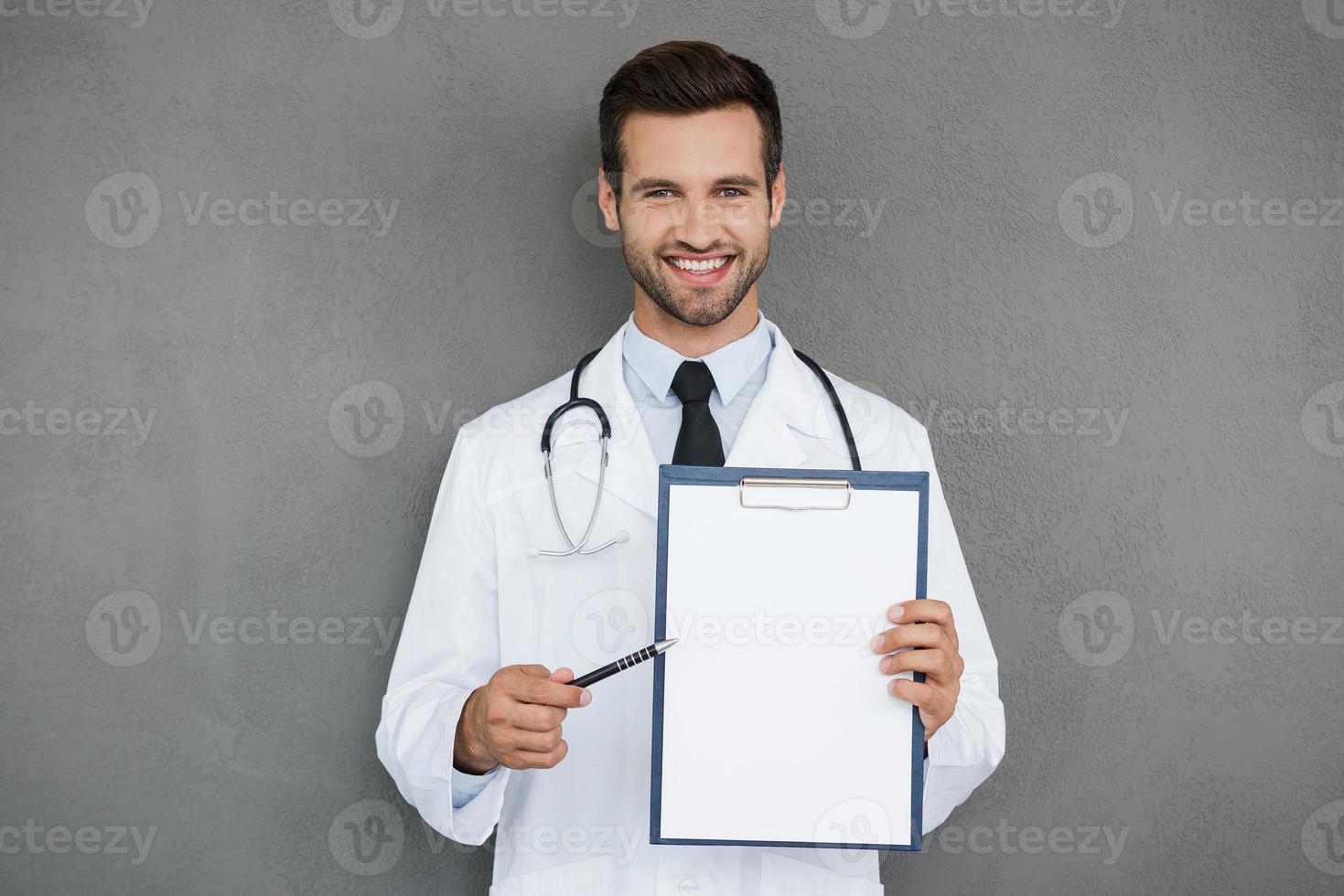 dokter richten kopiëren ruimte. glimlachen jong dokter in wit uniform Holding klembord en richten Bij het terwijl staand tegen grijs achtergrond foto