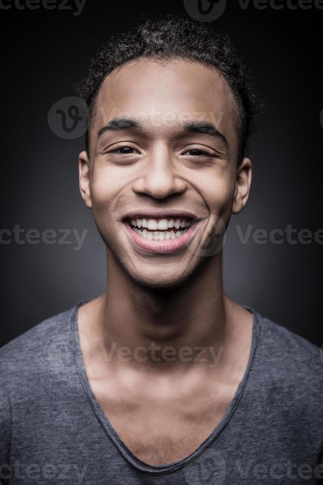 glimlach is mijn stijl. portret van vrolijk jong Afrikaanse Mens op zoek Bij camera met glimlach terwijl staand tegen zwart achtergrond foto