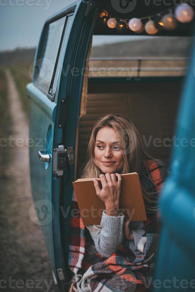 geïnspireerd door haar boek... aantrekkelijk jong vrouw gedekt met deken op zoek weg en glimlachen terwijl zittend binnen van de blauw retro stijl mini busje foto