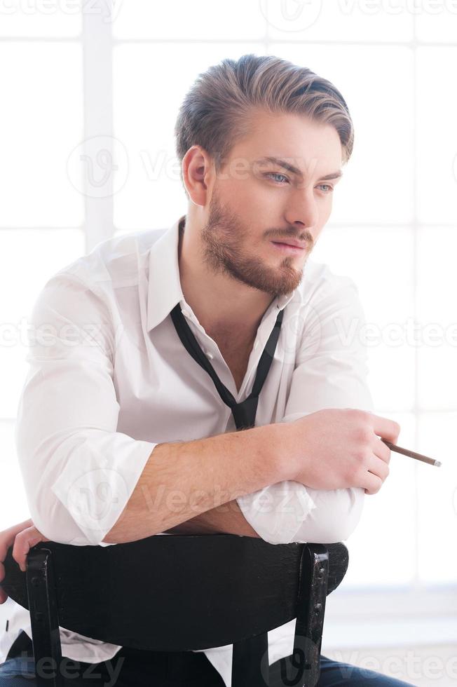 hebben een roken pauze. attent jong Mens in overhemd en stropdas roken een sigaret en op zoek weg terwijl zittend Aan de stoel foto