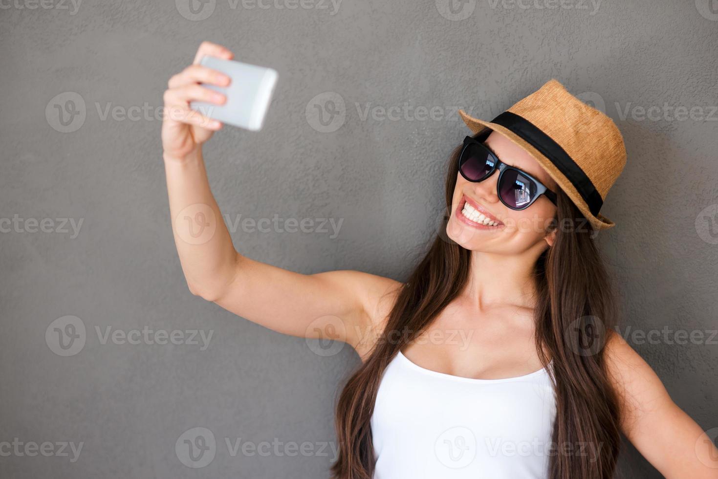 selfie tijd. blij jong Dames maken selfie door haar slim telefoon en glimlachen terwijl staand tegen grijs achtergrond foto