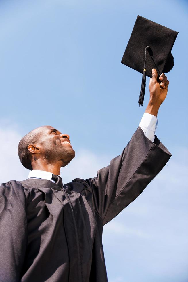 gelukkig naar worden afgestudeerd. laag hoek visie van gelukkig jong Afrikaanse Mens in diploma uitreiking japon Holding zijn Mortier bord tegen blauw lucht foto