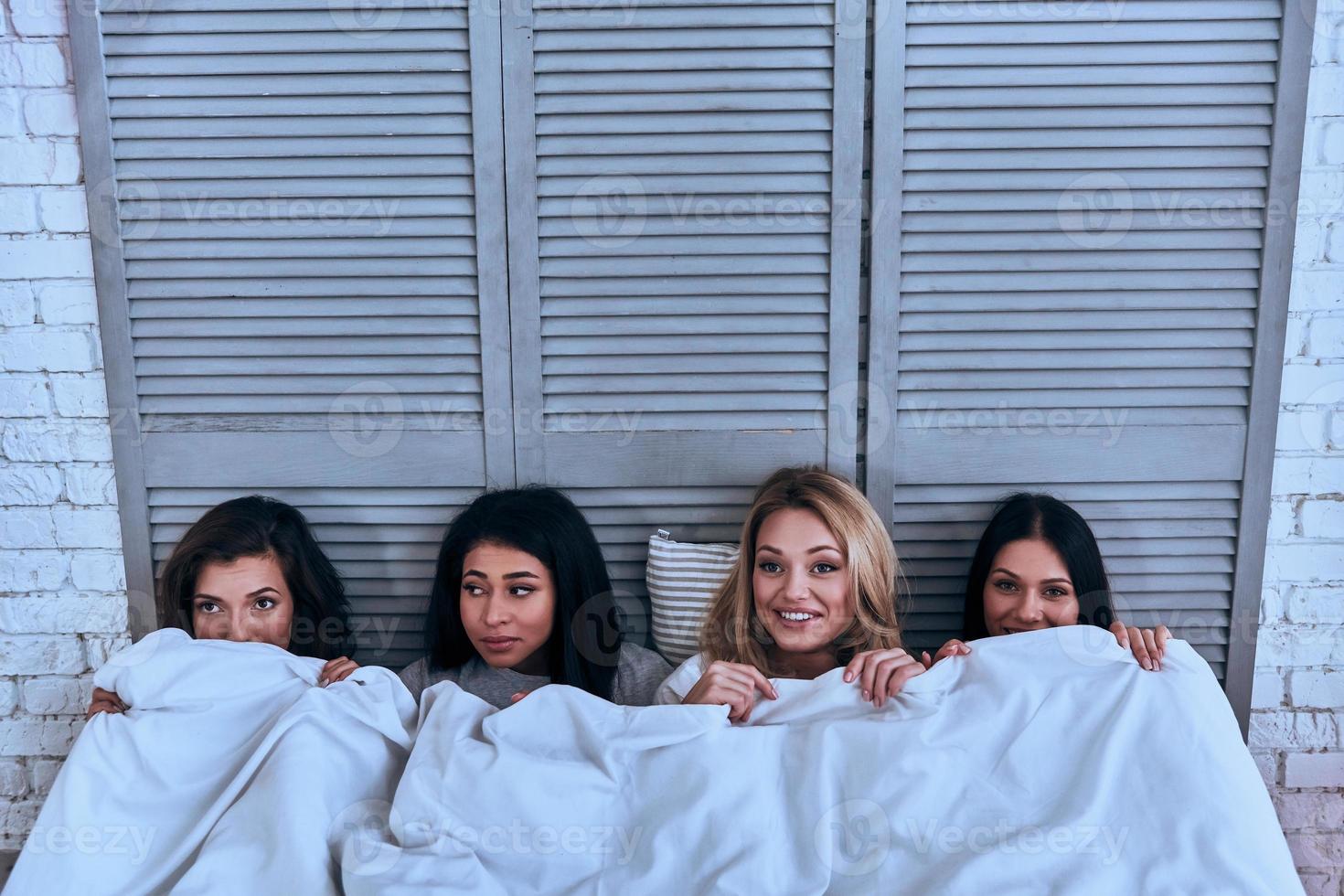 tijd naar slaap. top visie van vier mooi jong Dames aan het bedekken met wit deken en glimlachen terwijl aan het liegen in de bed foto