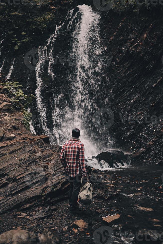 iedereen zou moeten zien het. vol lengte achterzijde visie van jong Mens op zoek Bij de waterval terwijl wandelen in de bergen foto