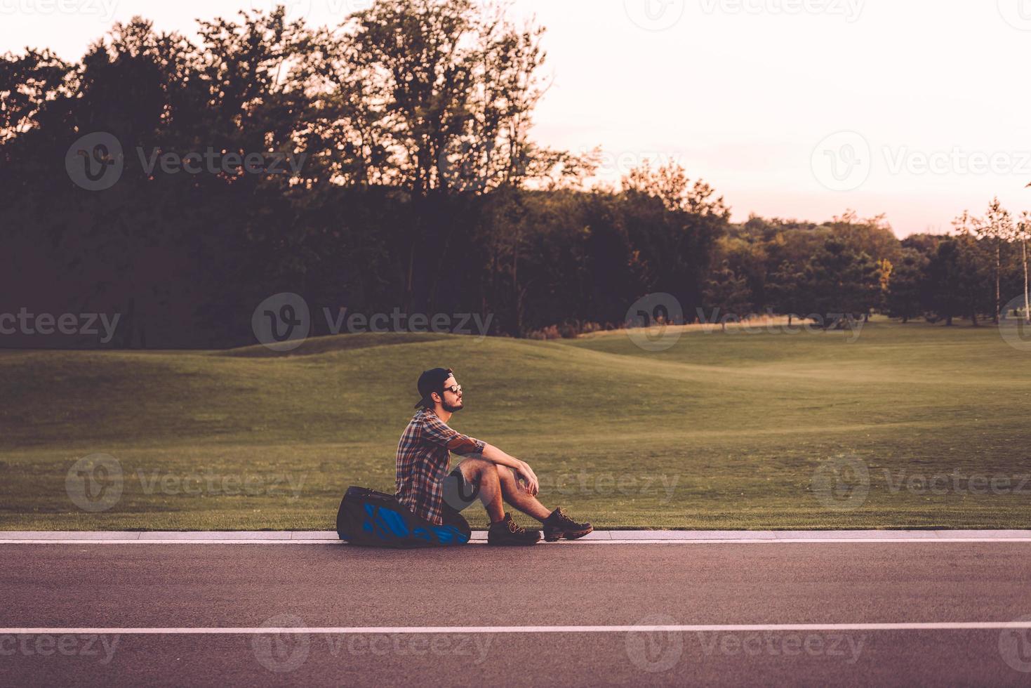 moe reiziger. kant visie van knap jong Mens zittend Aan zijn rugzak Bij de kant van de weg en op zoek attent foto