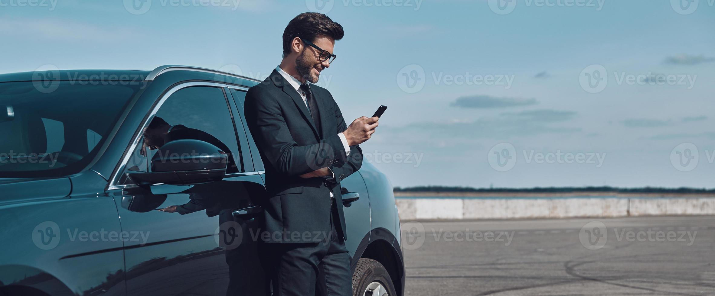 zelfverzekerd manager. vol lengte van knap jong zakenman gebruik makend van slim telefoon terwijl staand in de buurt zijn auto buitenshuis foto