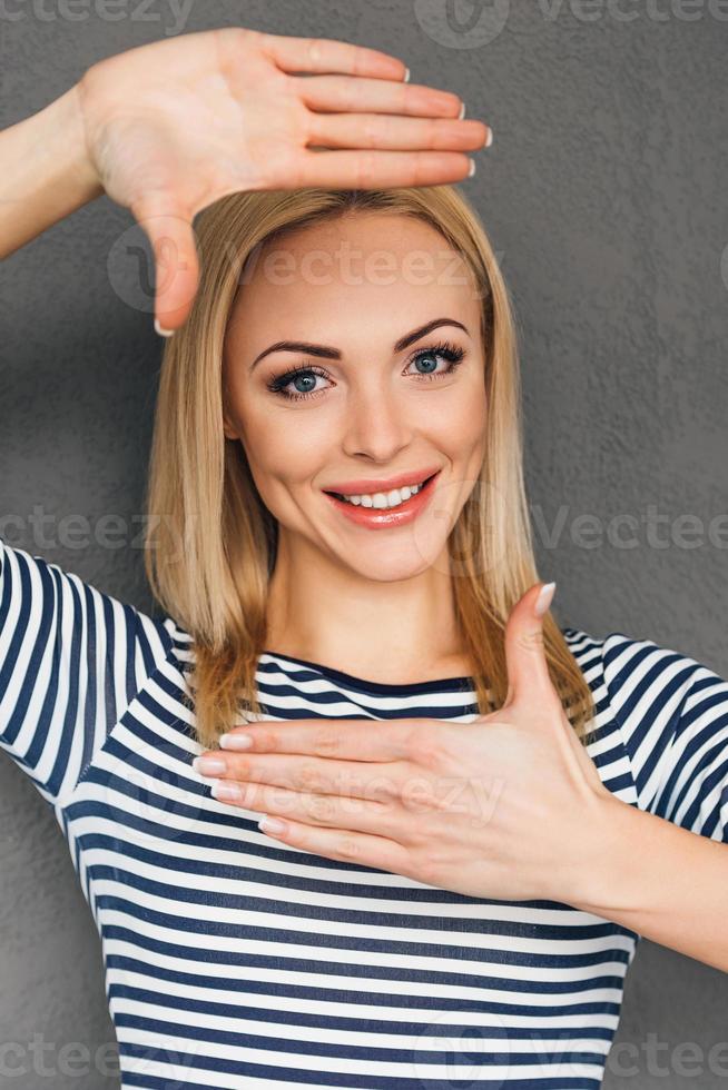 focus Aan me mooi jong vrouw maken vinger kader en op zoek Bij camera met glimlach terwijl staand tegen grijs achtergrond foto