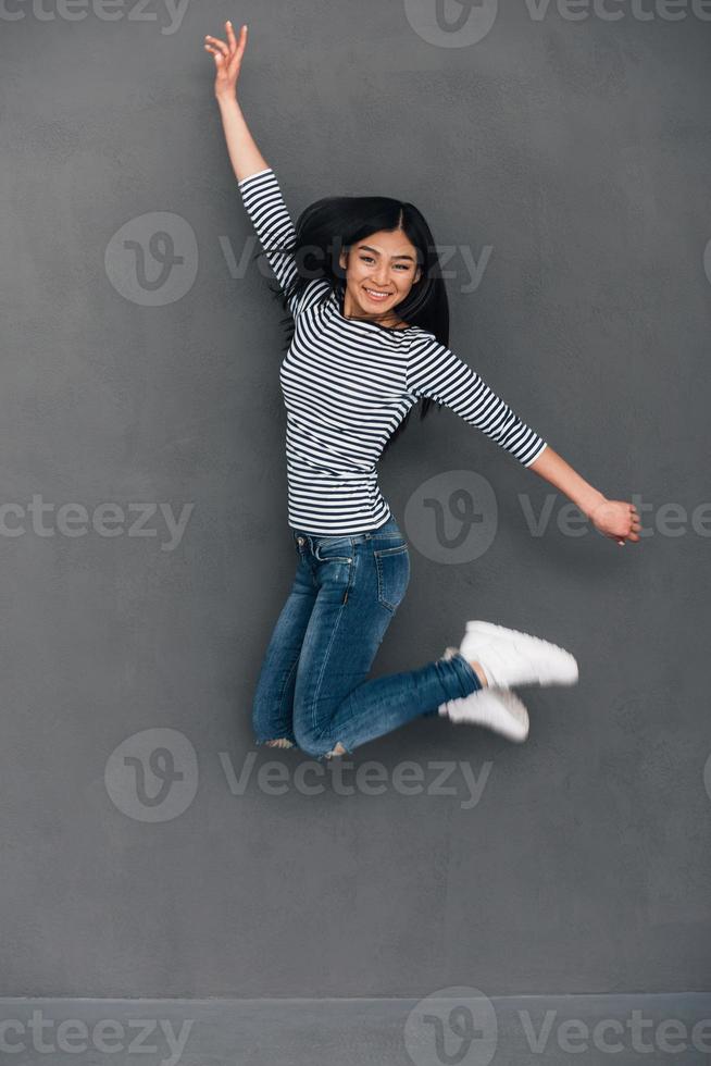 pret in in de lucht. mooi jong vrolijk Aziatisch vrouw op zoek Bij camera met glimlach terwijl jumping tegen grijs achtergrond foto