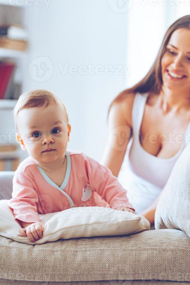 schattig en schattig. weinig baby meisje op zoek Bij camera en leunend Aan kussenterwijl haar moeder zittend Aan de bankstel Bij achtergrond foto