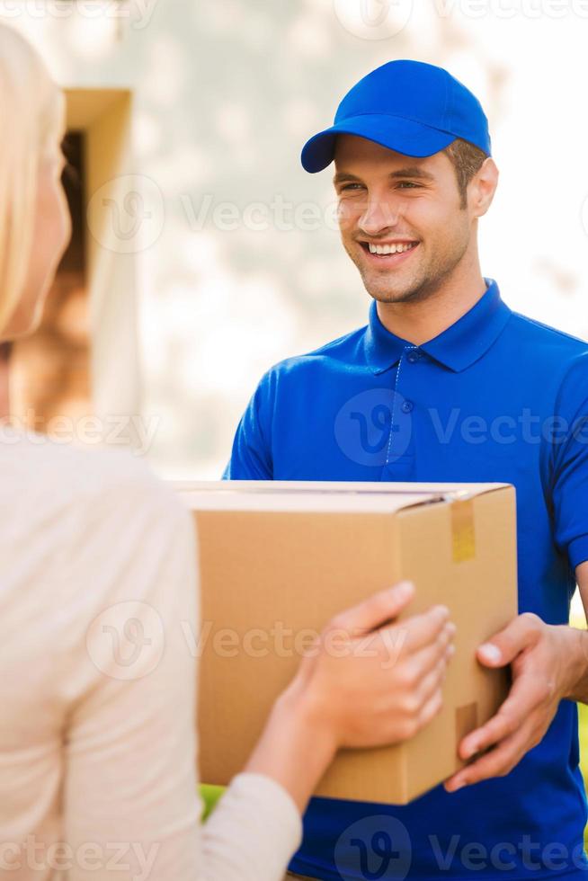 leveren elk pakket met glimlach. gelukkig jong koerier geven een karton doos naar jong vrouw terwijl staand buitenshuis foto