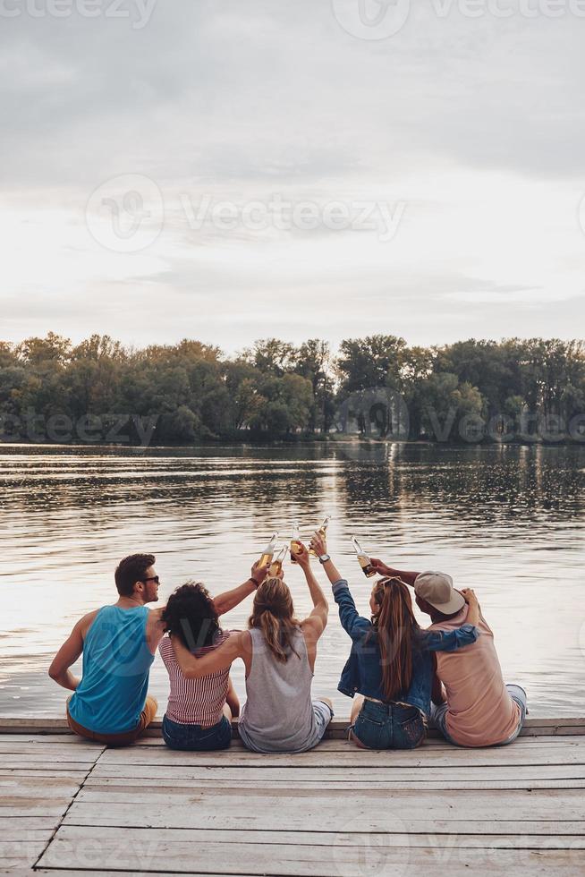 het beste reis ooit. achterzijde visie van jong mensen in gewoontjes slijtage roosteren met een bier flessen terwijl zittend Aan de pier foto