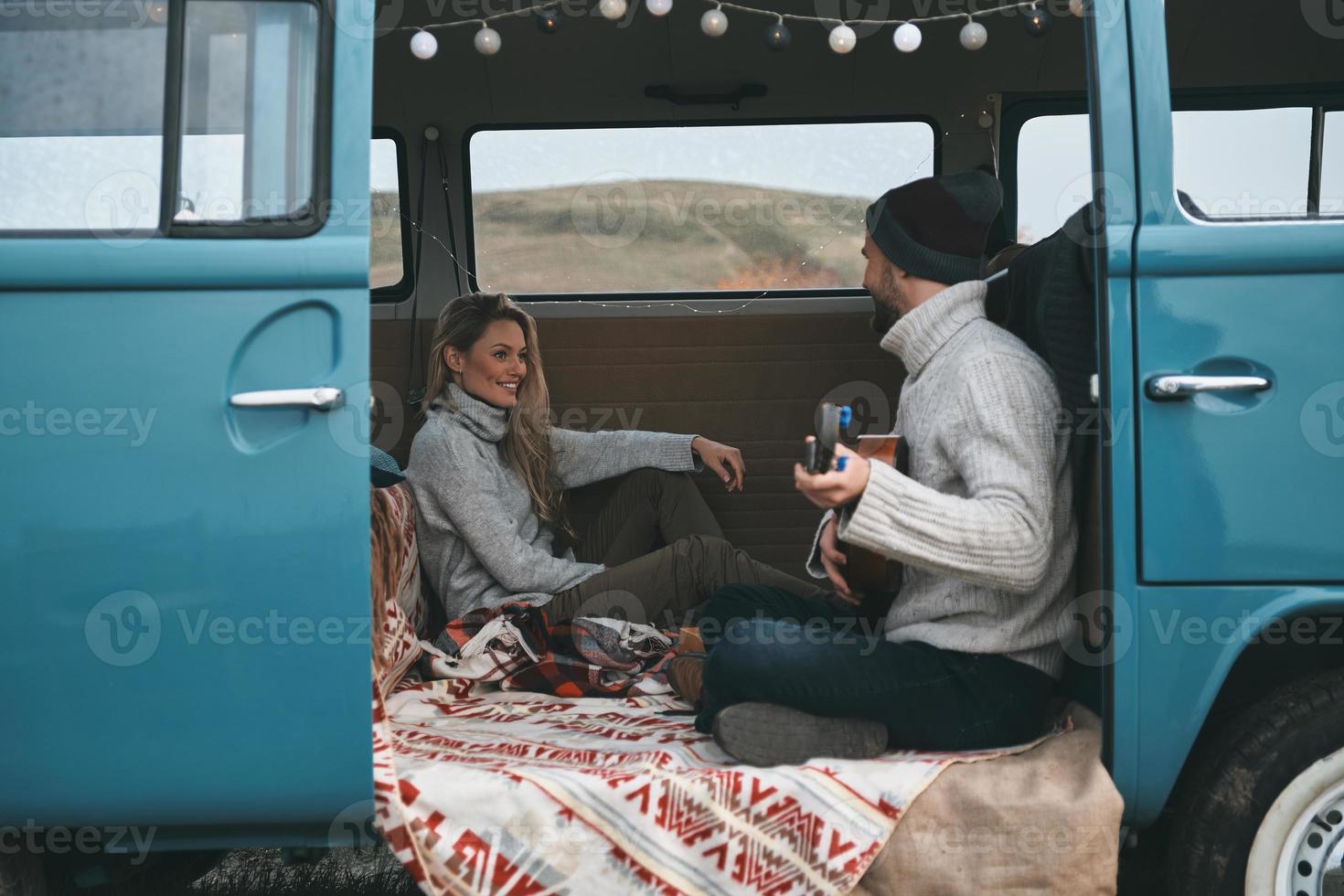 het zingen reizen liedje. knap jong Mens spelen gitaar voor zijn mooi vriendin terwijl zittend in blauw retro stijl mini busje foto