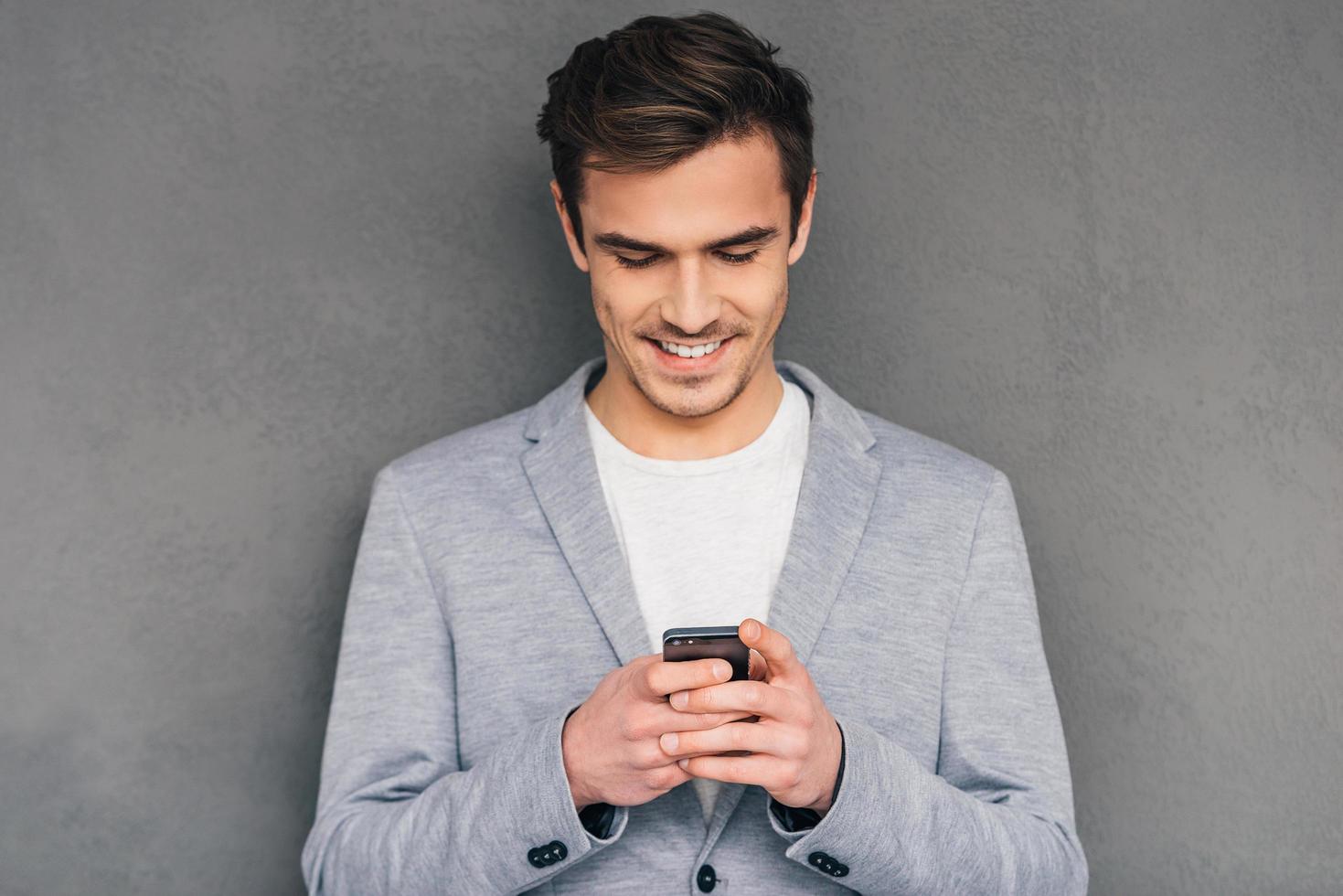 mooi zo bedrijf bericht. vrolijk jong Mens Holding mobiel telefoon en op zoek Bij het met glimlach terwijl staand tegen grijs achtergrond foto