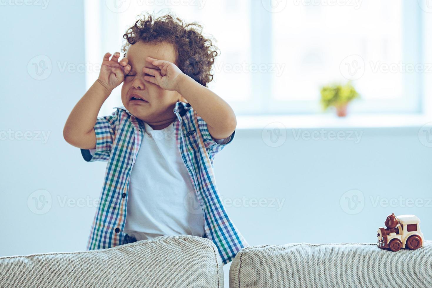 weinig Afrikaanse baby jongen huilen en wrijven zijn ogen terwijl staand Aan de bankstel Bij huis foto