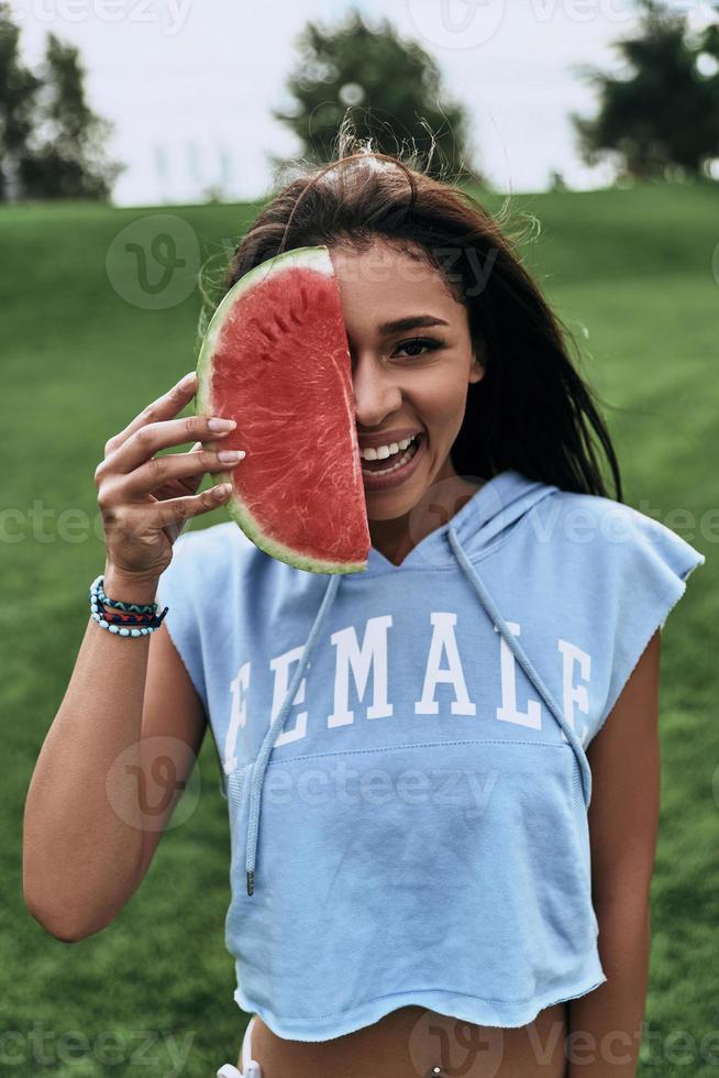 speels schoonheid. aantrekkelijk jong vrouw aan het bedekken een voor de helft van haar gezicht met een plak van watermeloen en glimlachen terwijl staand buitenshuis foto