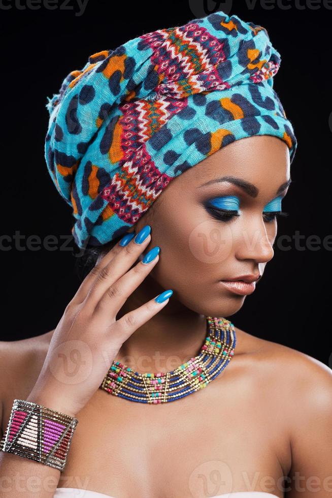 zelfverzekerd in haar perfect Look. mooi Afrikaanse vrouw vervelend een hoofddoek en ketting op zoek weg en aanraken haar gezicht terwijl staand tegen zwart achtergrond foto