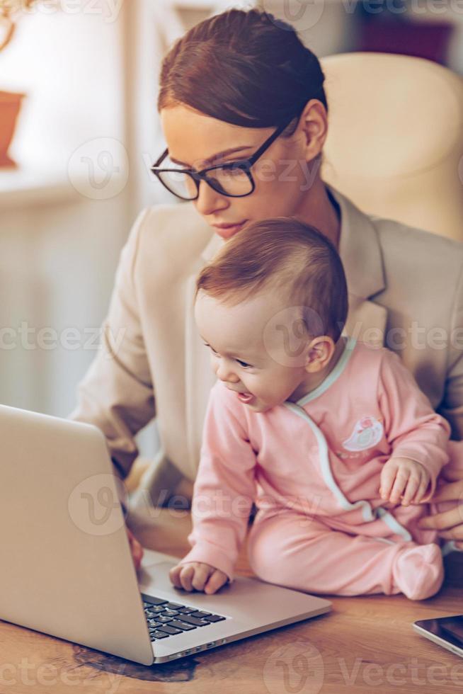 maken bedrijf plan met mama. weinig baby meisje gebruik makend van laptop terwijl zittend Aan kantoor bureau met haar moeder in kantoor foto