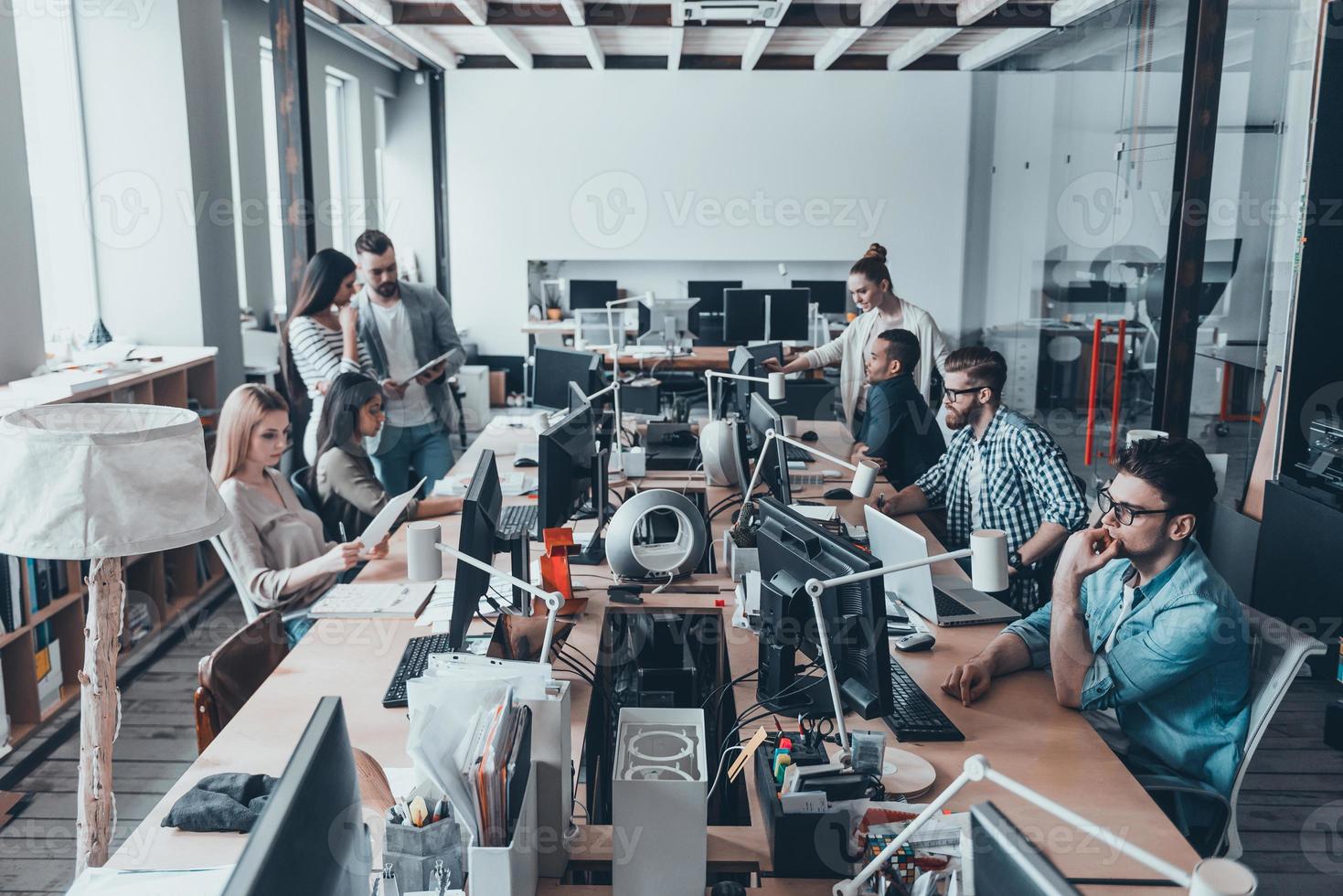 bezig werken dag in kantoor. groep van jong bedrijf mensen in slim gewoontjes slijtage werken en communiceren terwijl zittend Bij de groot bureau in de kantoor samen foto