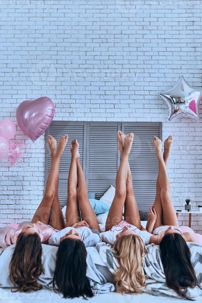 onweerstaanbaar meisjes. speels jong Dames houden voeten omhoog terwijl aan het liegen Aan de bed met ballonnen allemaal over- de kamer foto