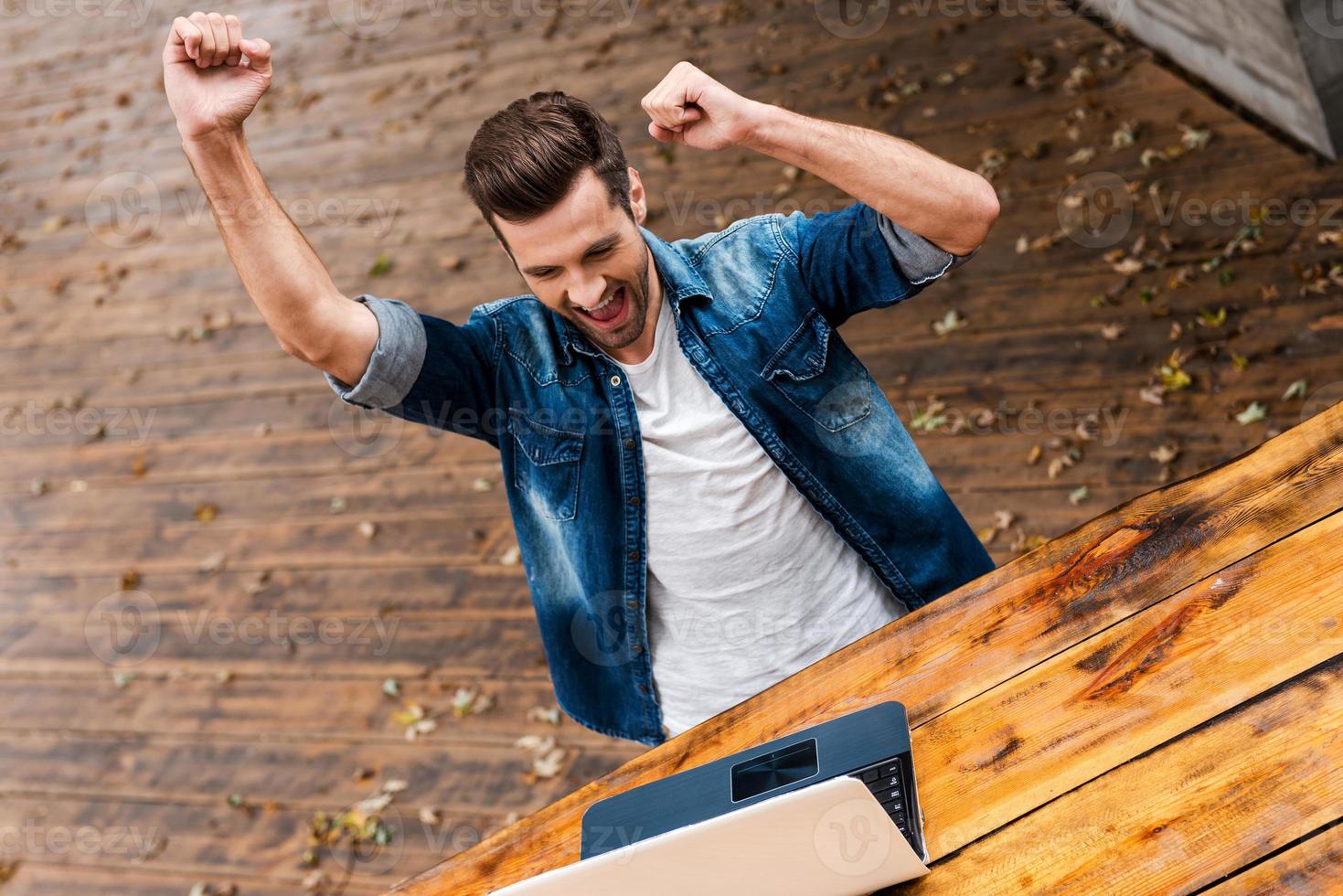 elke dag winnaar. top visie van opgewonden jong Mens houden armen verheven en uitdrukken positiviteit terwijl zittend Bij de houten tafel buitenshuis foto