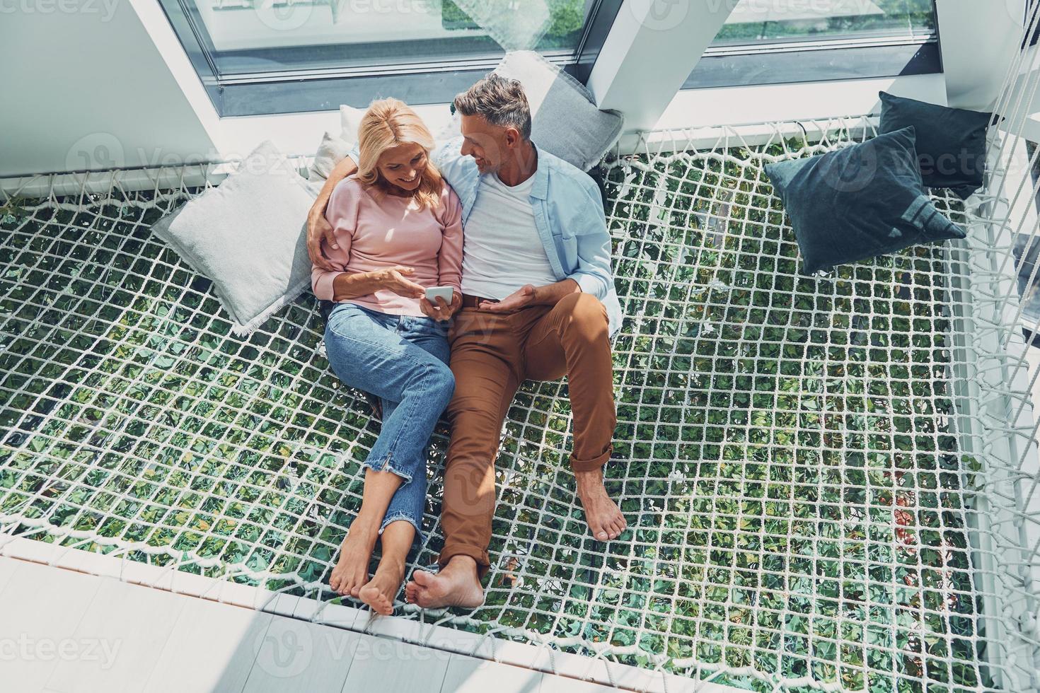 mooi volwassen paar onderzoeken slim telefoon en glimlachen terwijl ontspannende in groot hangmat Bij huis samen foto