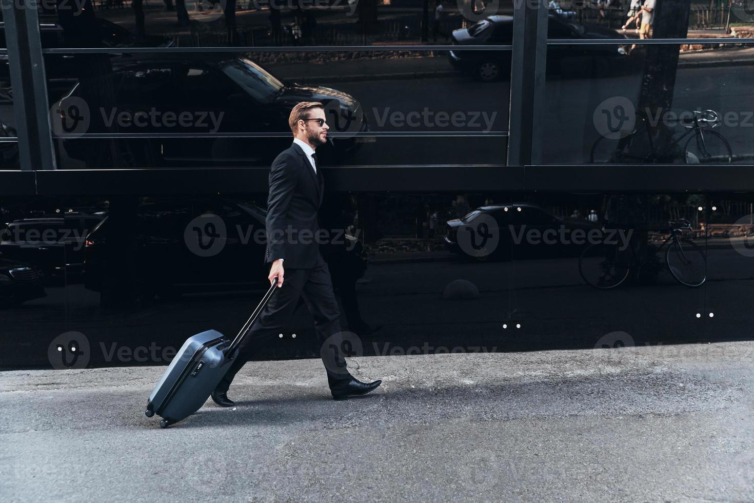 bedrijf reizen. vol lengte van jong Mens in vol pak trekken bagage terwijl wandelen buitenshuis foto