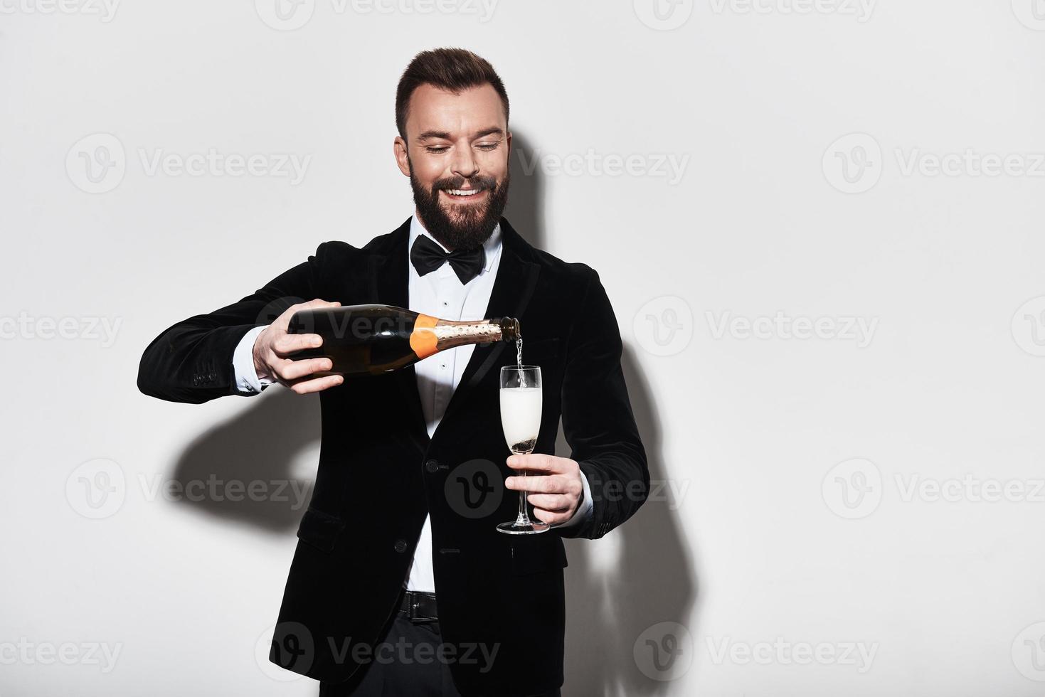 klaar naar feest. knap jong Mens in vol pak gieten Champagne en glimlachen terwijl staand tegen grijs achtergrond foto